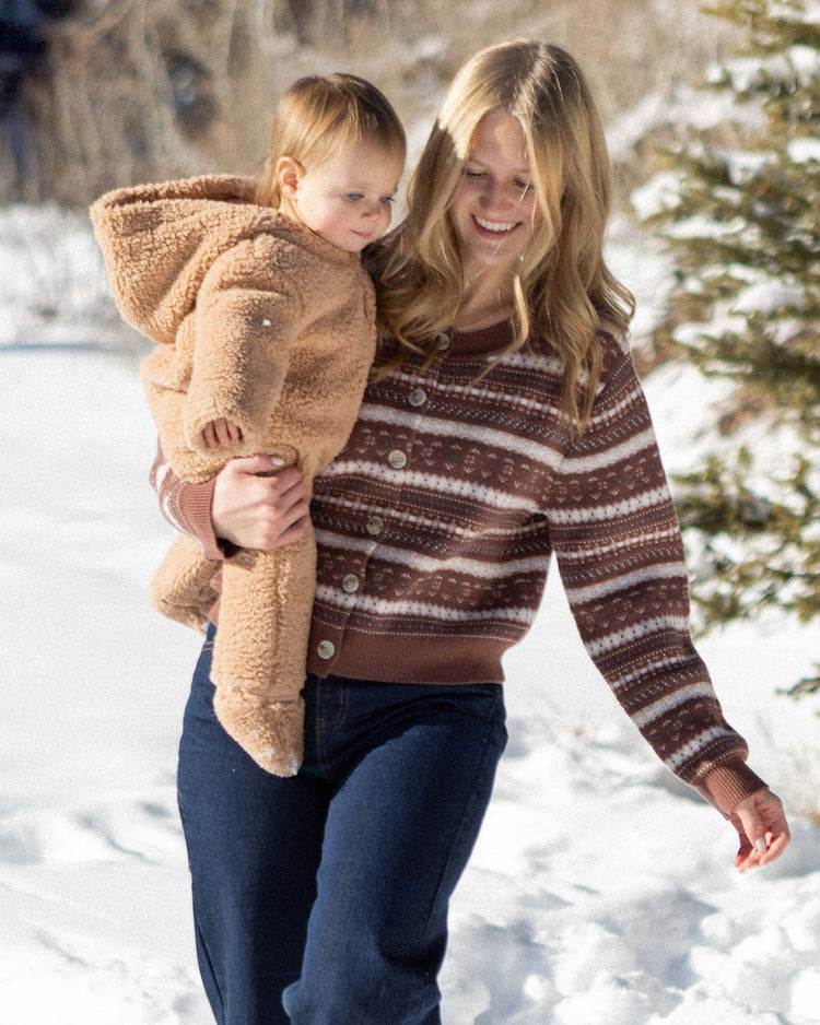A woman in a striped sweater smiles while holding a baby wearing the rylee + cru fleece jumpsuit in fawn. They are walking outside in the snow near some pine trees.