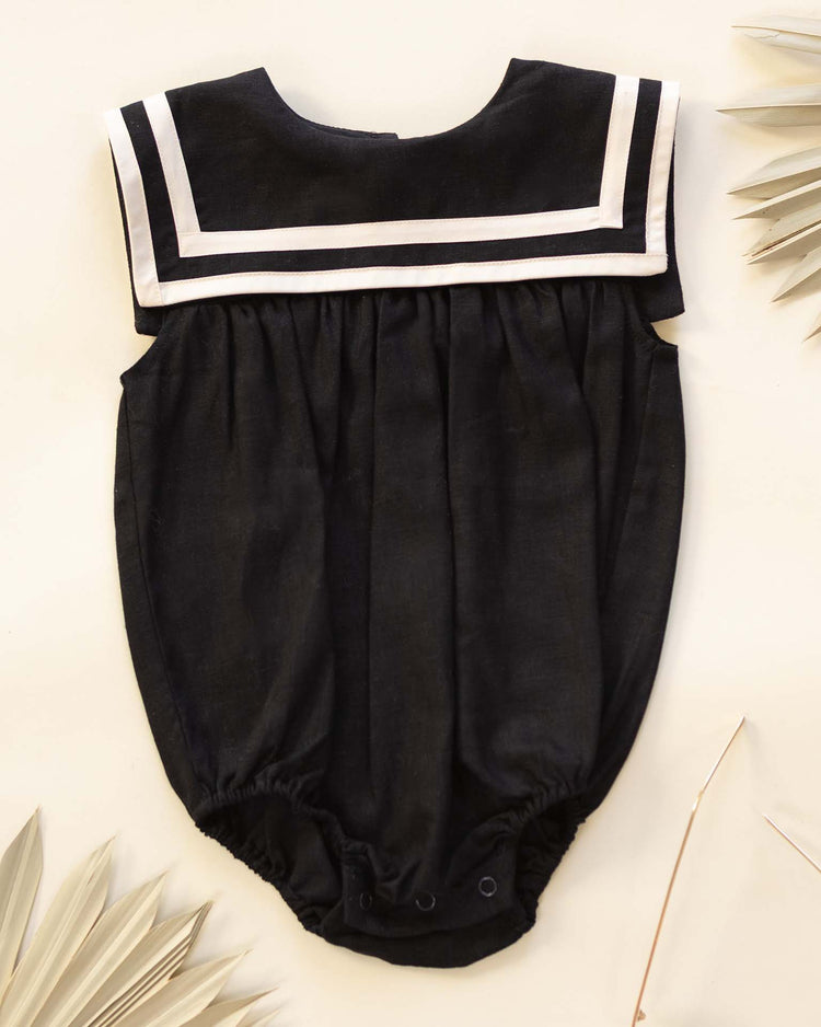 The rylee + cru sailor romper in black features a sleeveless design and white-trimmed sailor collar, shown laid flat on a light surface with dried leaves next to it.