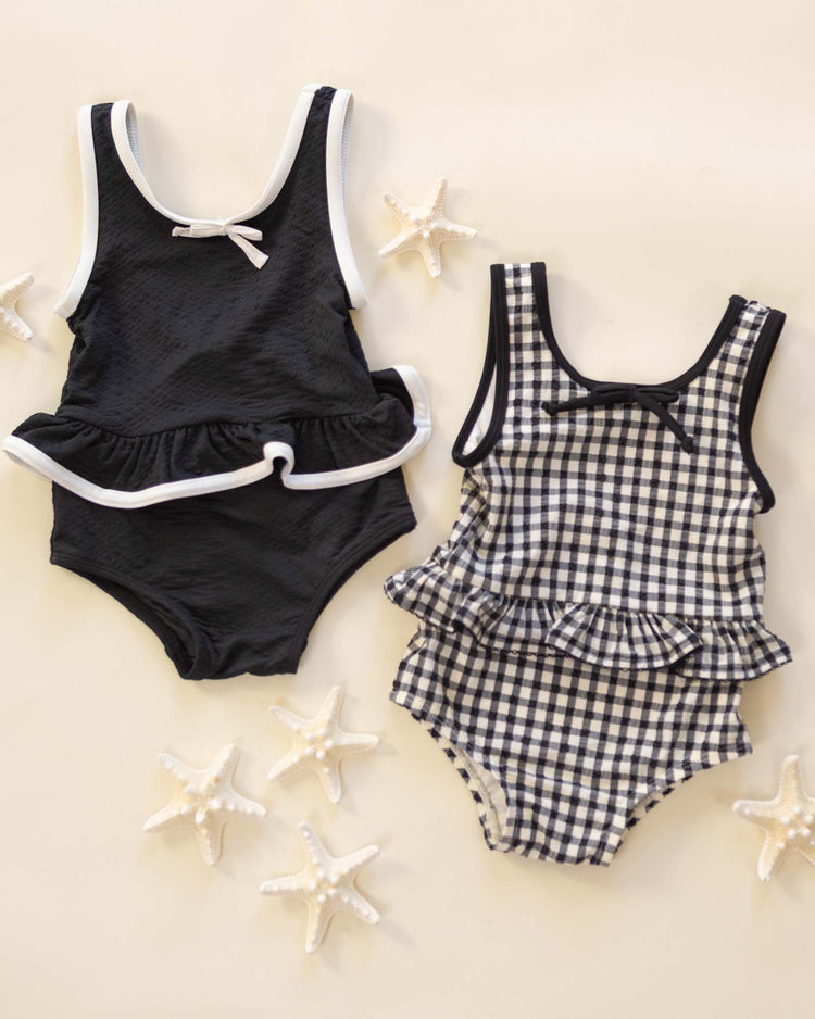 Two rylee + cru children's swimsuits lay flat on a light surface with six starfish: one is a solid black skirted one-piece with white trim and ruffle, the other is a black gingham skirted one-piece with ruffle and bow.