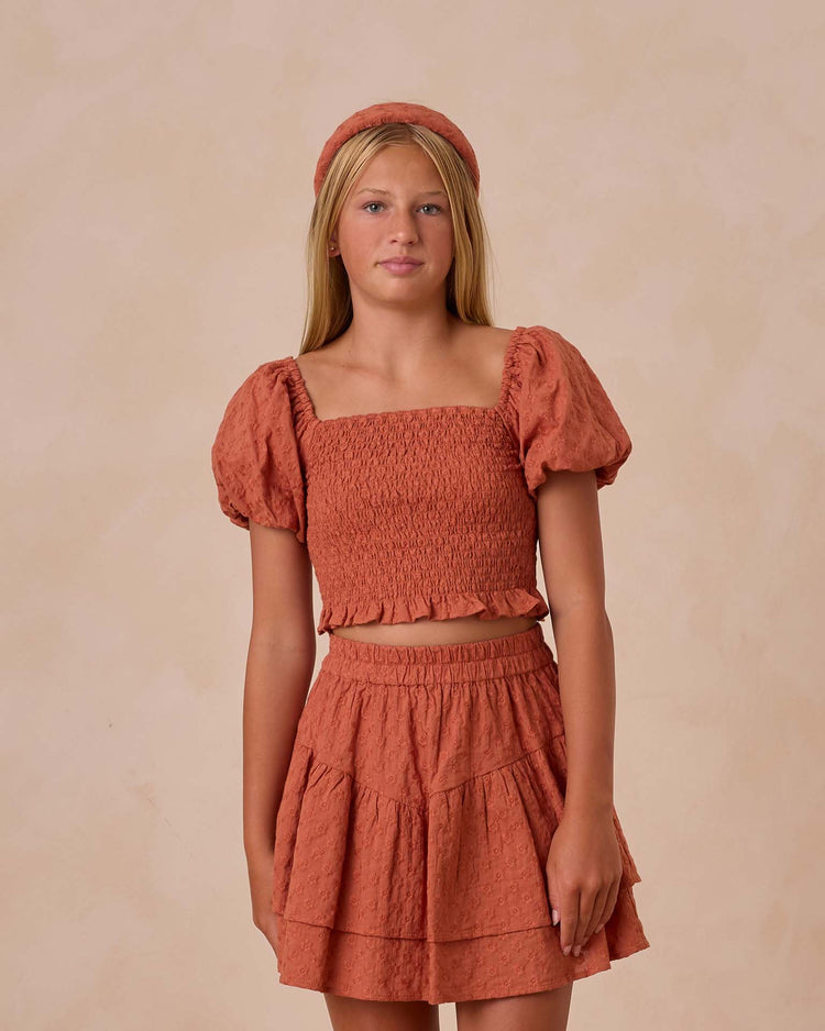 A young girl in the Rylee + Cru teen trixie top in poppy, with bubbled sleeves and a tiered skirt, stands against a beige background.