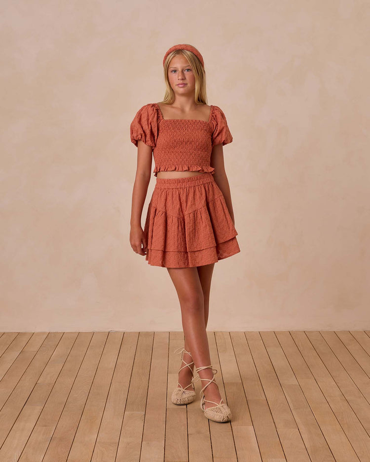 A person stands on a wooden floor wearing the rust-colored teen trixie top in poppy by rylee + cru with bubbled sleeves and a smocked bodice, styled with a matching skirt and headband against a neutral background.