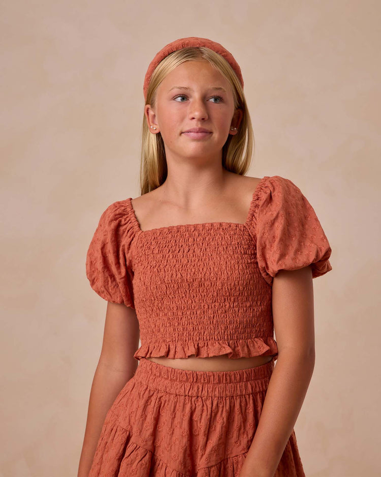 A young girl with long blonde hair wears the teen trixie top in poppy by rylee + cru, featuring a smocked bodice and bubbled sleeves with a matching headband, against a plain background.