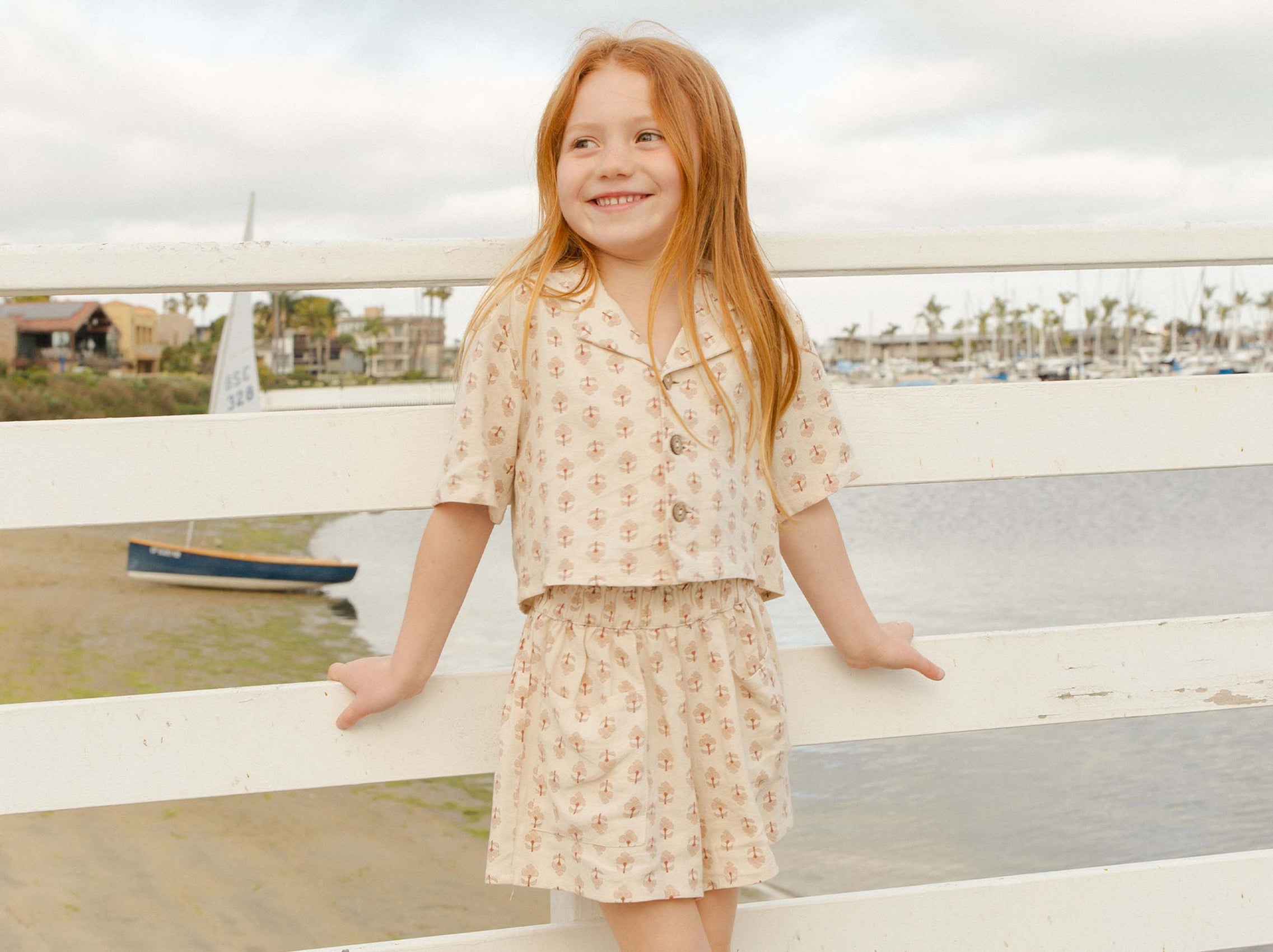 Young girl with red hair smiling and leaning on a white fence by a marina with boats and overcast sky in the background.
