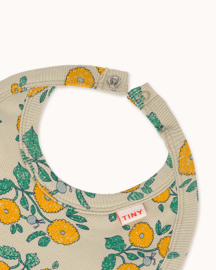Baby bib named "flowers bib in dark vanilla" by tiny cottons, showcasing a yellow and green floral design on a light beige background with a snap closure labeled "TINY.