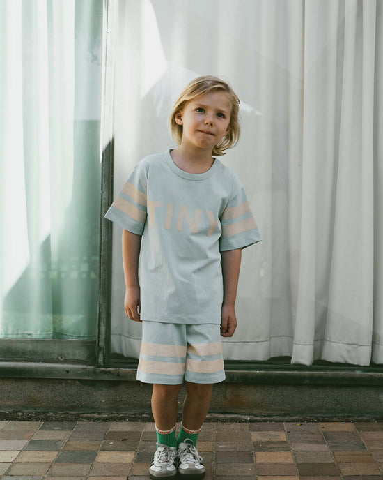 A child stands on a brick path wearing a light-colored shirt and stripes logo short in dusty blue from tiny cottons, paired with green socks and sneakers. White curtains hang gracefully in the background, adding an airy touch to the ensemble.