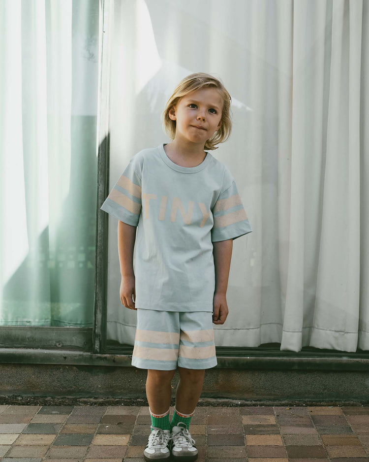 A child stands on a brick path wearing a light-colored shirt and stripes logo short in dusty blue from tiny cottons, paired with green socks and sneakers. White curtains hang gracefully in the background, adding an airy touch to the ensemble.
