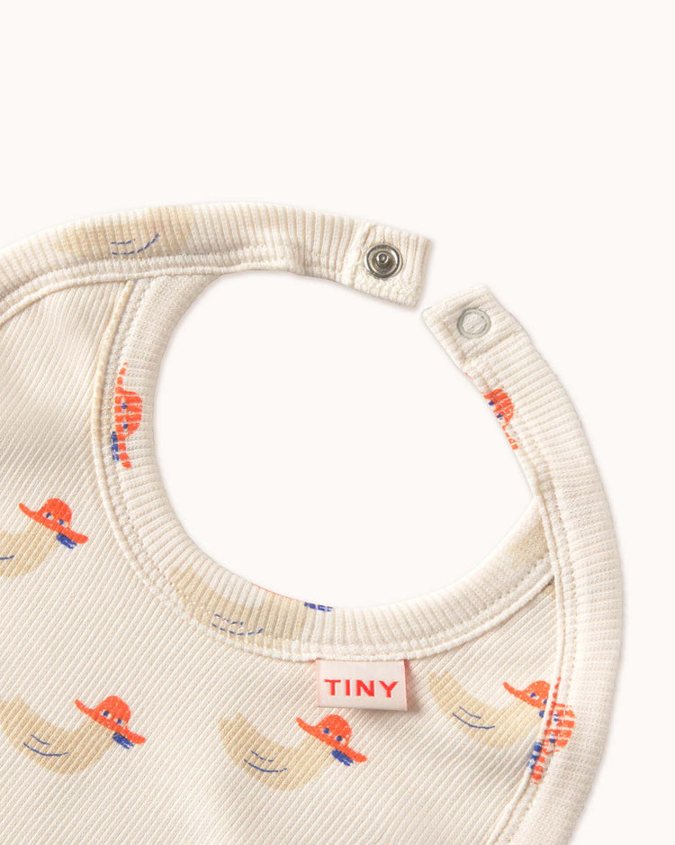 The "ducks bib in off-white" by Tiny Cottons is a high-quality baby bib made from organic cotton, with colorful elephants and the word "TINY" on a label, featuring a snap closure on the top edge against a plain background.