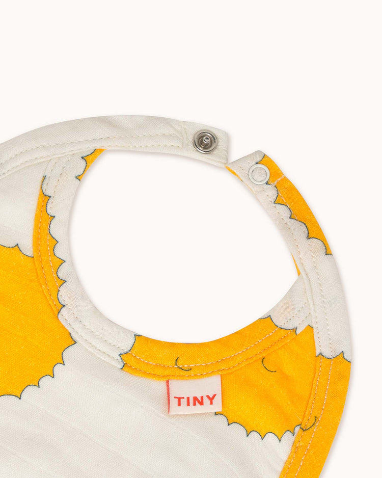 A close-up of the sun bib in off-white reveals a yellow and white pattern with a snap closure. Made from soft organic cotton by tiny cottons, the label highlights unique prints adding whimsy.