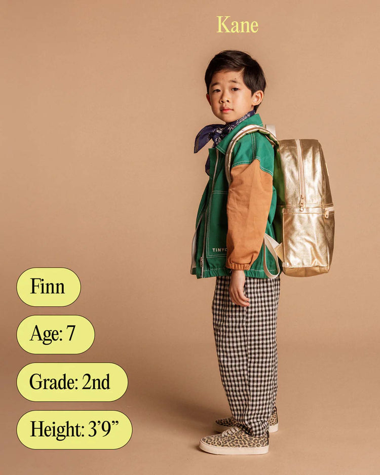 A young boy stands sideways wearing a green and tan jacket, checkered pants, a blue scarf, and a gold Kane backpack in Happy Fuzzy Hearts by STATE Bags. Text: Kane above, and labels: Finn, Age: 7, Grade: 2nd, Height: 3’9”.