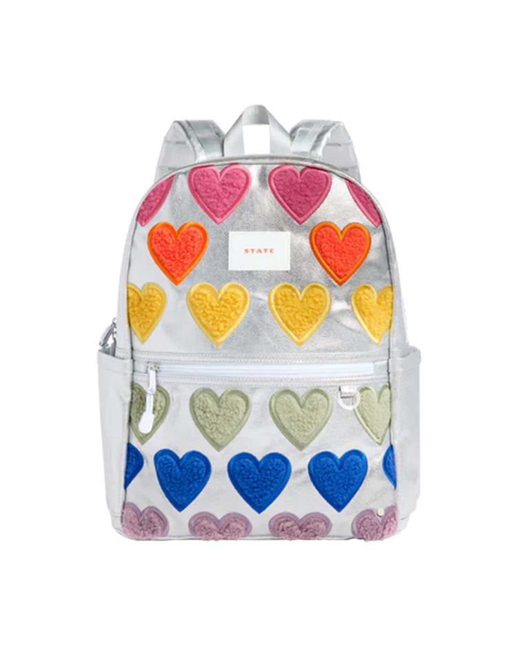Introducing the Kane Backpack in Happy Fuzzy Hearts by STATE Bags—a silver travel backpack adorned with rows of colorful heart patches in red, yellow, green, blue, and purple. This stylish school backpack features a front zipper pocket, convenient side pockets, and eye-catching metallic shoulder straps.
