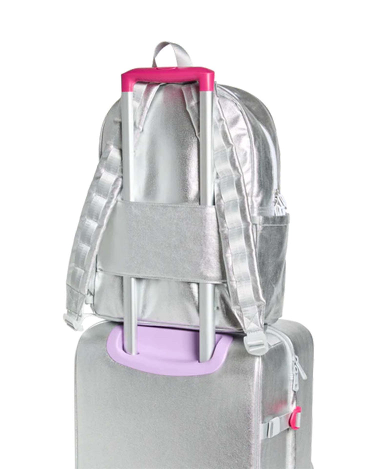 A State Bags Kane Backpack in Happy Fuzzy Hearts, featuring silver with pink and lavender accents, is secured to the handle of a matching silver rolling suitcase. This eye-catching travel backpack by State Bags stands out beautifully against the white background.