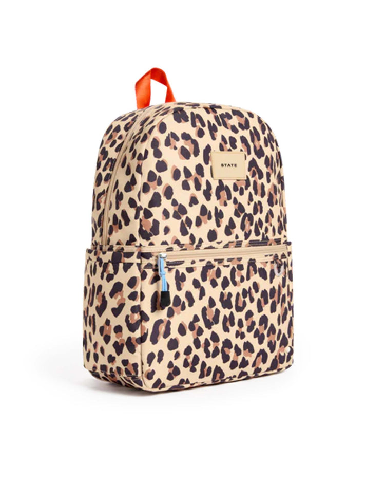 The Kane backpack in leopard by State Bags features a tan base with a brown and black leopard print, a front zipper pocket, side pockets, and an orange top handle. This stylish backpack is displayed against a plain white background.