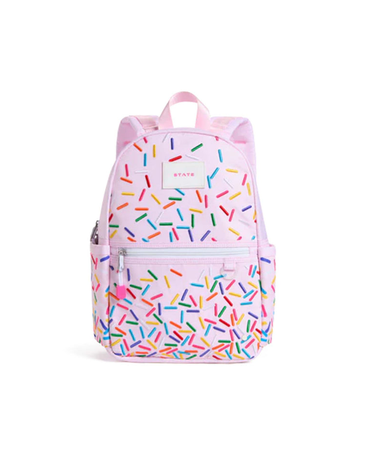 The kane mini backpack in sprinkles by state bags is a light pink kids backpack decorated with colorful sprinkle patterns. It features a front zipper pocket, side pockets, a top handle, and the "STATE" brand label displayed on the front.