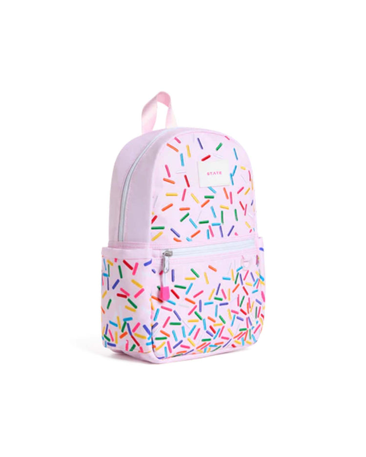 The kane mini backpack in sprinkles by state bags features a playful pastel pink design adorned with colorful sprinkle patterns, a front zipper pocket, convenient side pockets, and a pink carrying handle—making it the perfect kids backpack for everyday adventures.