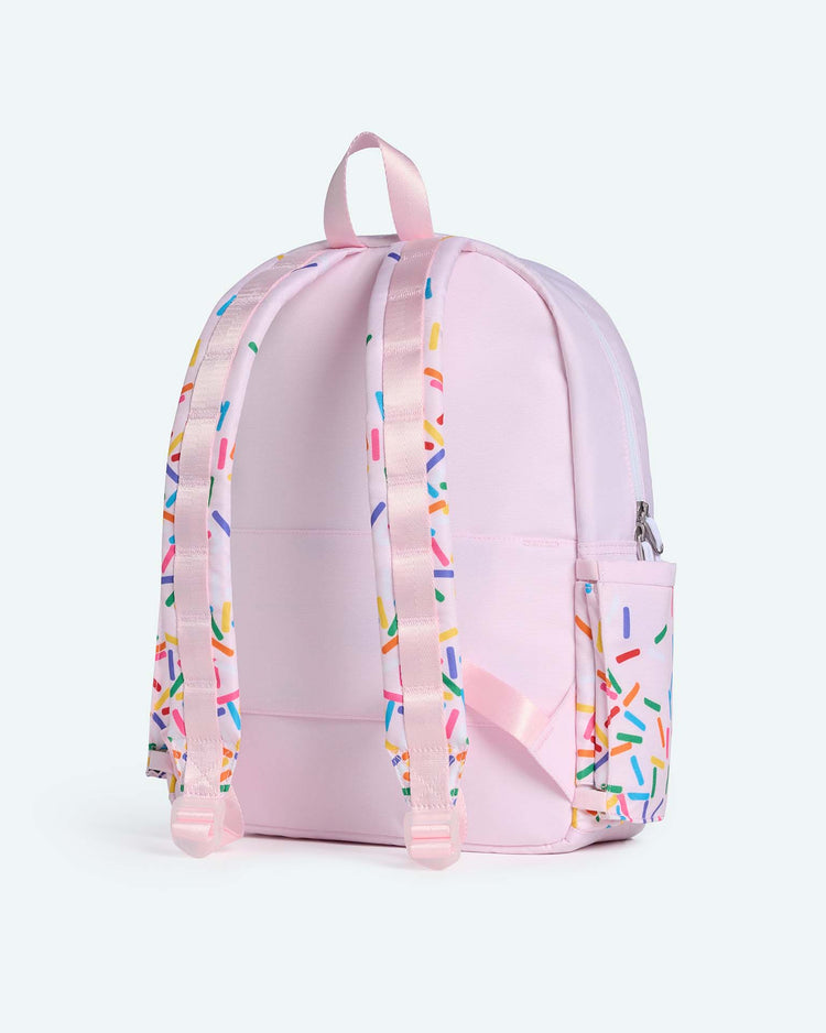 The state bags kane backpack in sprinkles features a pastel pink design adorned with colorful sprinkle patterns on the straps and side pocket, showcased against a plain light blue background. Inspired by the classic Kane travel backpack, this playful accessory is perfect for adding a pop of fun to your everyday adventures.