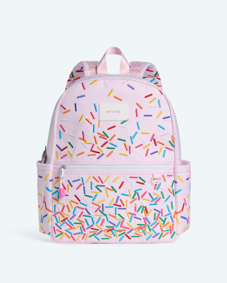 The Kane Backpack in Sprinkles by STATE Bags is a pink school backpack adorned with colorful sprinkle patterns, featuring a front zipper pocket, side pockets, and a small STATE Bags label near the top center.