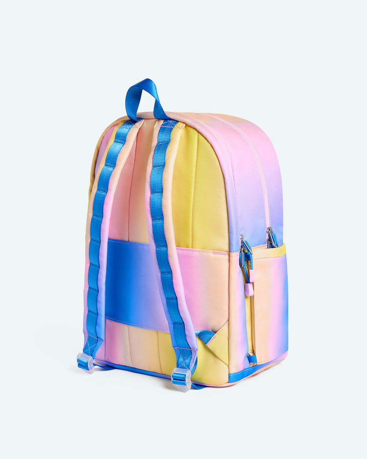 Introducing the Kane Large Double Pocket Backpack from State Bags, featuring a blue, pink, and yellow gradient design. This stylish backpack is equipped with blue adjustable straps, a top handle, convenient side pockets, and a secure zipper closure. The soft blend of pastel colors makes it an ideal choice for school-goers who want both functionality and standout style.