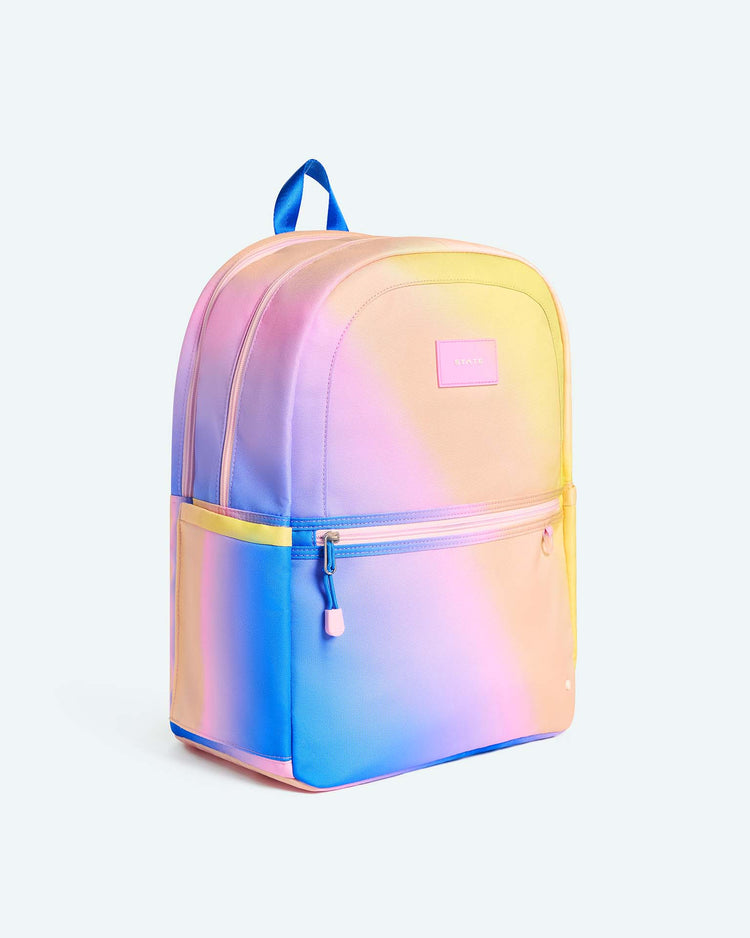 A kane large double pocket backpack by state bags, showcasing a blue, pink, and yellow gradient with front and side pockets plus a blue handle, is displayed on a light background—perfect as a stylish school backpack.
