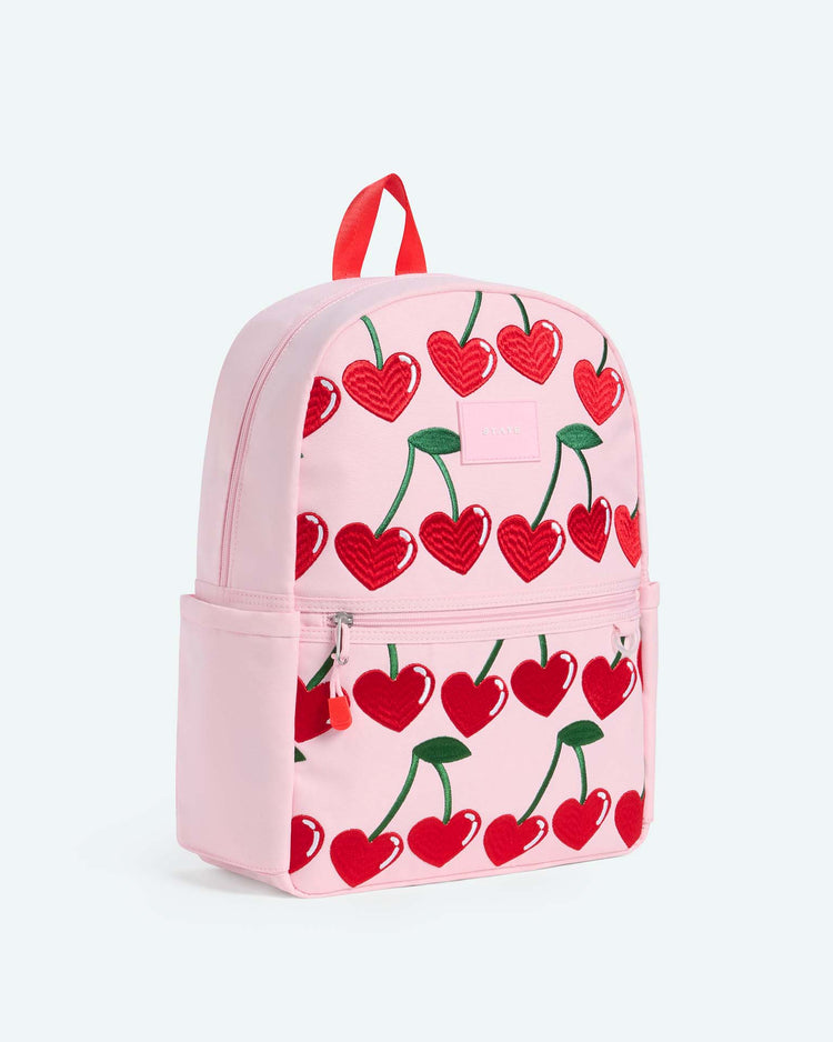 Introducing the kane backpack in cherry by state bags—a light pink travel backpack featuring bold red heart-shaped cherry prints, a handy front pocket, two side pockets, and a striking red top handle. Perfect as a stylish school backpack or for weekend adventures.