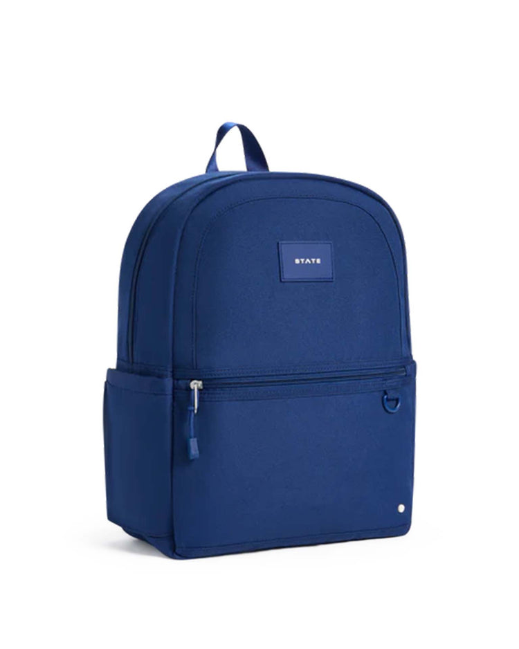 A STATE Bags Kane Backpack in navy, featuring a front zipper pocket, a convenient side pocket, and a sturdy top handle. The front is adorned with a small rectangular label reading "STATE." Ideal for use as a school or travel backpack, it is displayed standing upright against a white background.