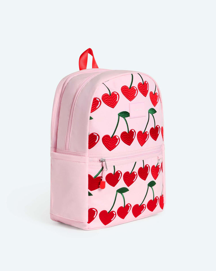 The Kane Double Pocket Backpack in Cherry by State Bags is a pink school backpack adorned with a red cherry pattern and green stems. It features a front zip pocket, a red zipper pull, and a matching red carrying loop on top.