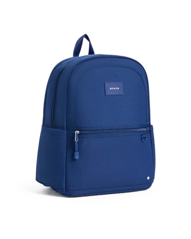 The Kane Double Pocket Backpack in Navy by State Bags features a solid blue design with front zippered pockets, a side pocket, and a small rectangular State Bags label on the front. The school backpack is shown standing upright against a white background.