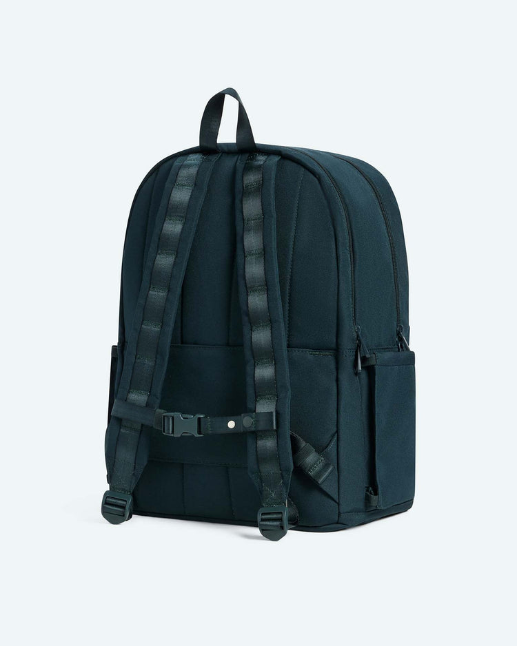 The state bags reed backpack in dark green is displayed from the back against a plain light background, highlighting its padded shoulder straps, chest strap, top handle, and convenient side pockets. This durable backpack is ideal for everyday use or as a dependable school companion.