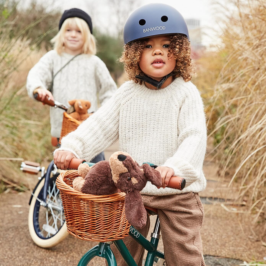 A child wearing a helmet and a white sweater stands with a teddy bear in a bicycle basket, while another child looks on from behind.