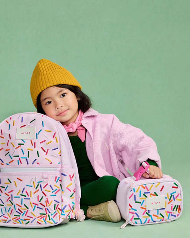 A young child in a yellow beanie and pink jacket sits against a green background, holding a matching pastel mini Rodgers snack pack in sprinkles by State Bags, decorated with colorful sprinkles.