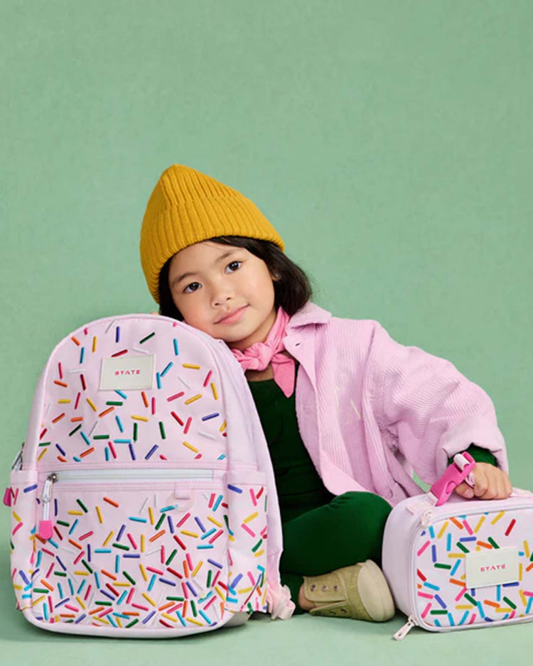 A young child in a yellow beanie and pink jacket sits on the floor, smiling, next to the STATE Bags Kane Mini Backpack in Sprinkles.