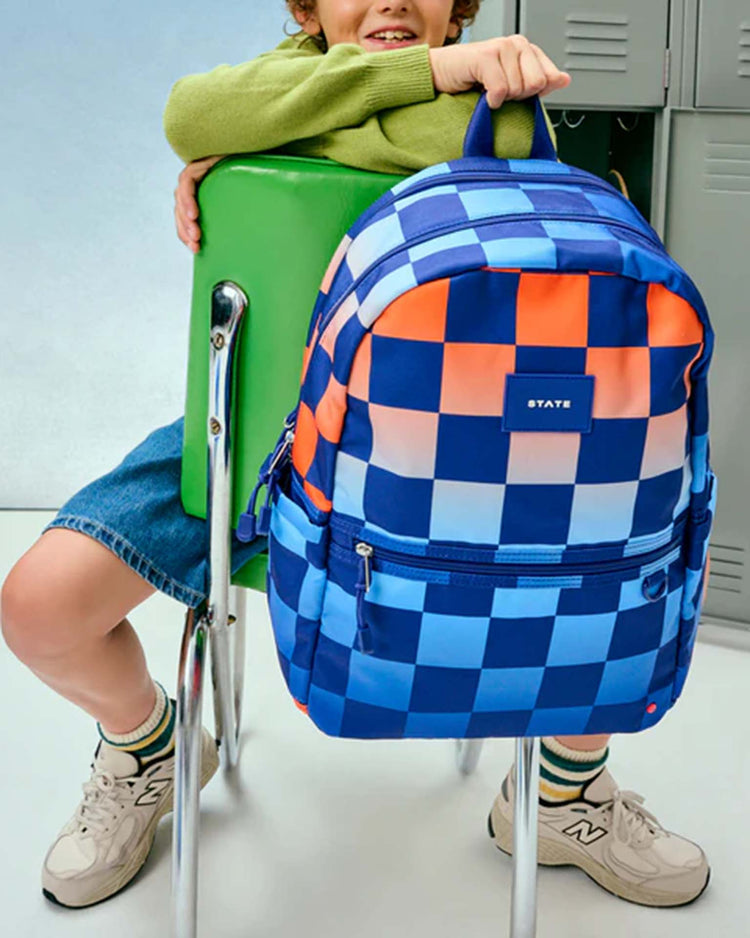 A child sits backward on a green chair, resting their arms on its back. Hanging from the chair is a State Bags Kane Double Pocket Backpack in Gradient Checkerboard, with school lockers visible in the background.