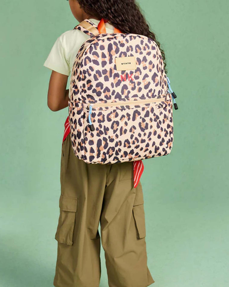 A person with long curly hair, dressed in a light shirt and olive cargo pants, stands facing away while carrying the State Bags Kane Backpack in Leopard against a plain mint-green background.