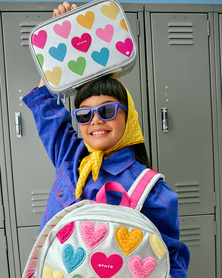 A smiling child in sunglasses and a yellow scarf holds the STATE Bags Rodgers lunch box in Happy Fuzzy Hearts above their head, wearing a matching kids’ lunch box backpack, standing in front of gray lockers.