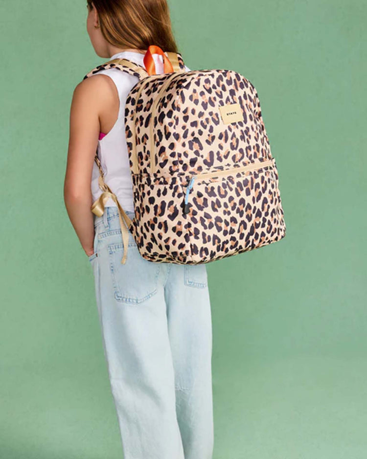 A young person with long hair, dressed in a white sleeveless top and light blue jeans, stands against a plain green background carrying the State Bags Kane Double Pocket Backpack in Leopard, which has a water bottle tucked into its side pocket.