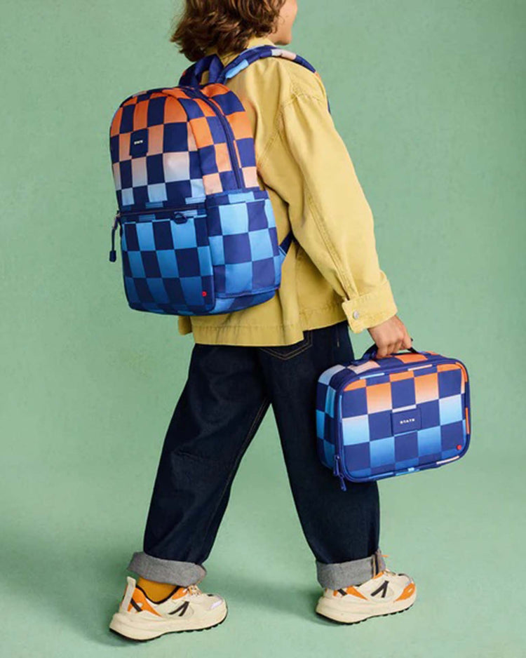 A child wearing a yellow jacket, dark jeans, and white sneakers is holding the STATE Bags rodgers lunch box in gradient checkerboard and a matching backpack, standing against a green background.