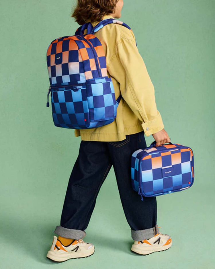 A child wearing a yellow shirt, blue jeans, and sneakers carries the STATE Bags Kane backpack in gradient checkerboard along with a matching lunchbox against a green background.