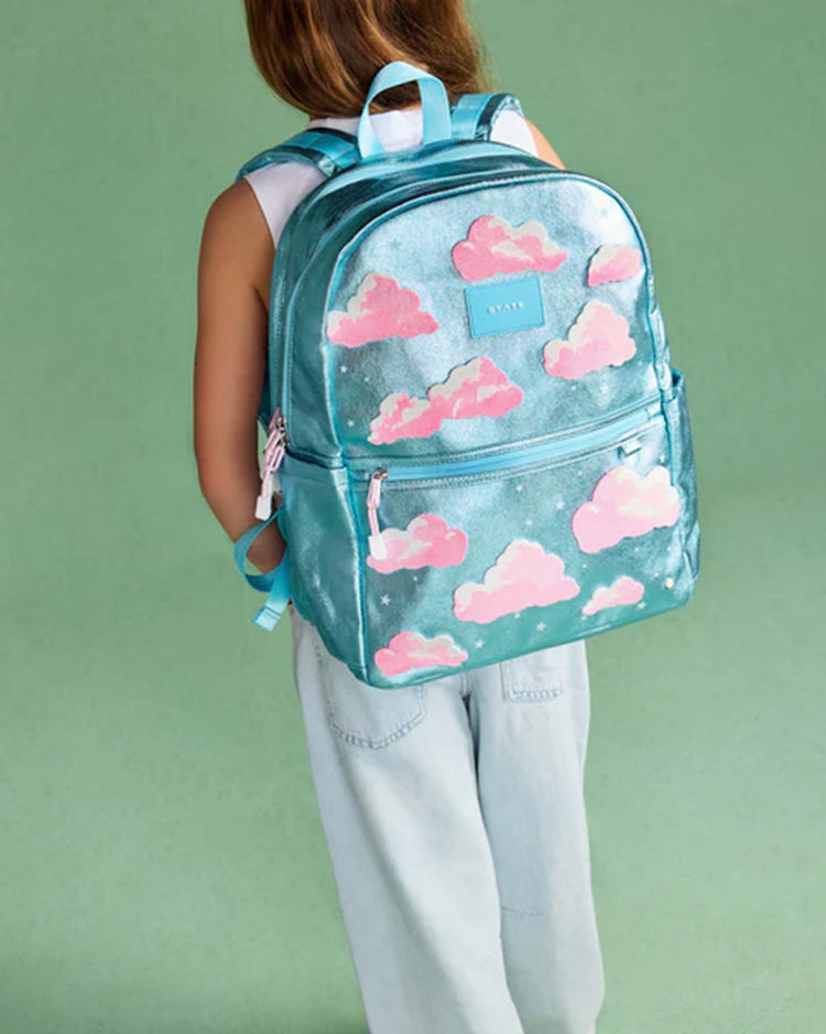 A child wearing a State Bags Kane Double Pocket Backpack in Clouds stands against a green background, facing away from the camera.