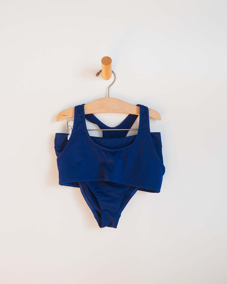 The sunchild luisa two-piece swimsuit in obscur features a tank-style top and high-waisted bottoms, displayed on a wooden hanger against a white wall—an ideal choice for girls' swimwear collections.