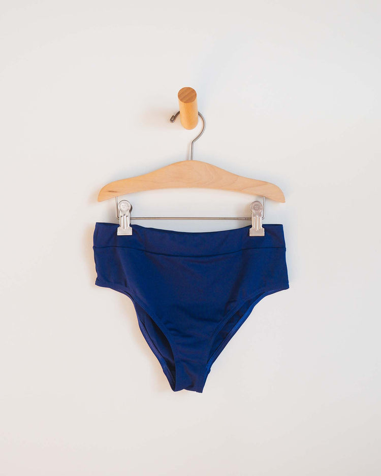 A pair of high-waisted, dark blue Sunchild luisa two-piece swimsuit bottoms in obscur, clipped to a wooden hanger and hanging on a peg against a plain white wall.