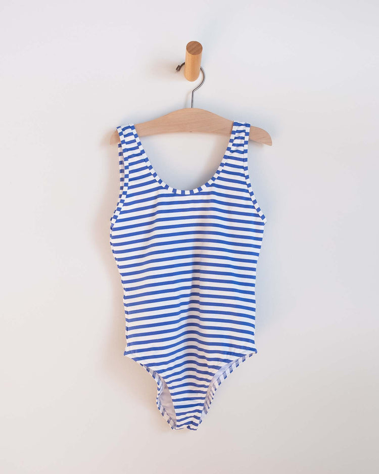 The sunchild lou one-piece swimsuit in mare, featuring blue and white stripes, hangs on a wooden hanger against a plain white wall—perfect for girls' swimwear collections.