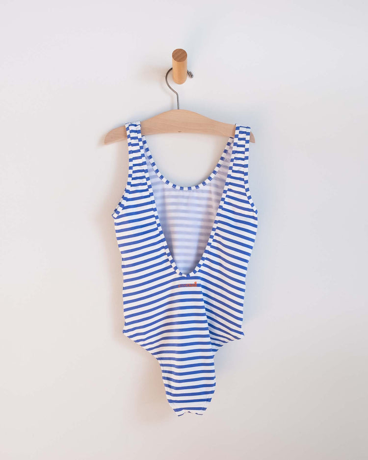 The sunchild lou one-piece swimsuit in mare, featuring blue and white stripes, hangs on a wooden hanger against a plain white wall—an ideal addition to any girl's swimwear collection.