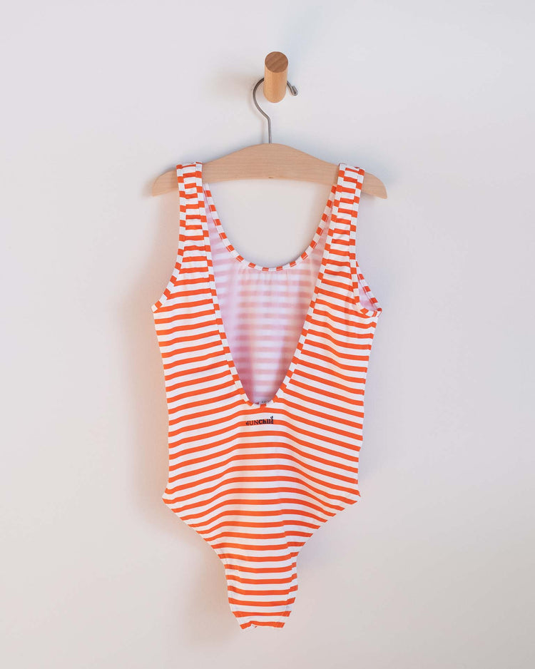 The sunchild lou one-piece swimsuit in soda, featuring red and white stripes and a deep V-neck, hangs on a wooden hanger from a wall-mounted peg against a plain white background.