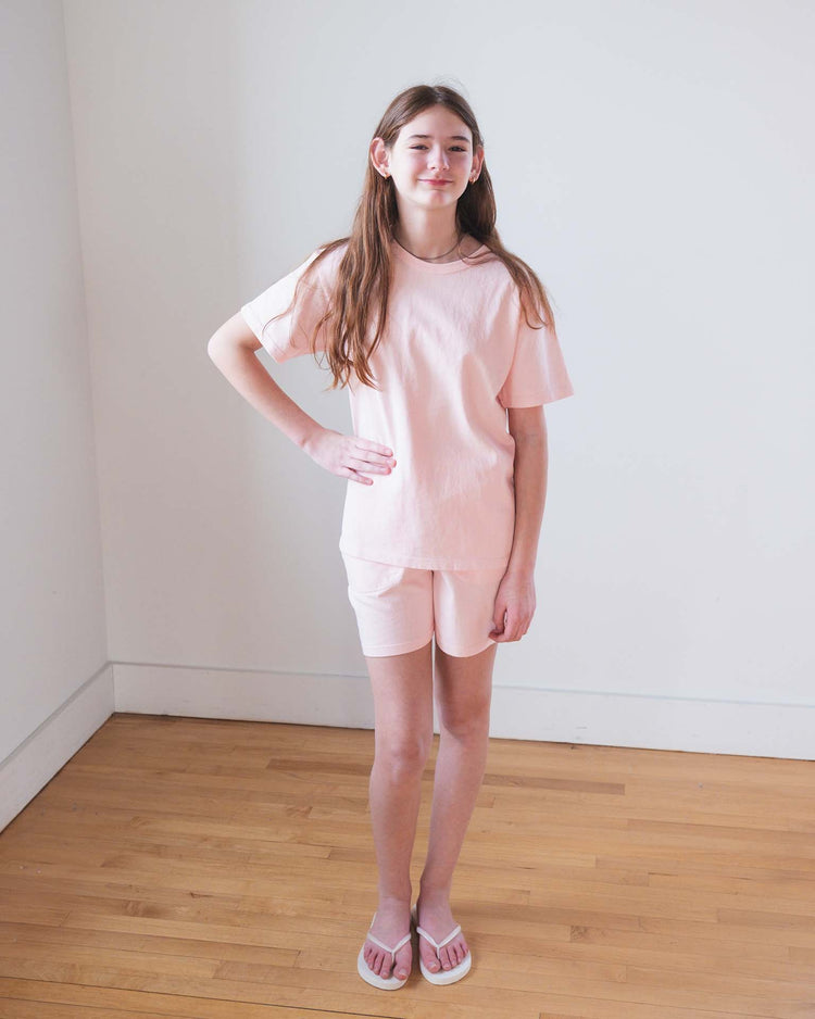 A young girl with long brown hair wears Sunchild's merida shorts in baby and a light pink t-shirt. She poses indoors on a wood floor, one hand on her hip, against a plain white wall, paired with white flip-flops.