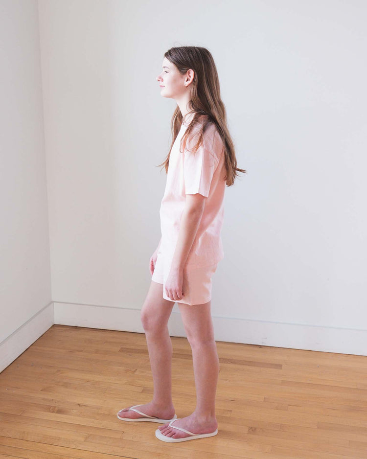 A young woman with long brown hair stands sideways in a bright, empty room wearing the Sunchild kazan tee in baby, matching shorts, and pink slippers on a wooden floor. She gazes ahead with a neutral expression.