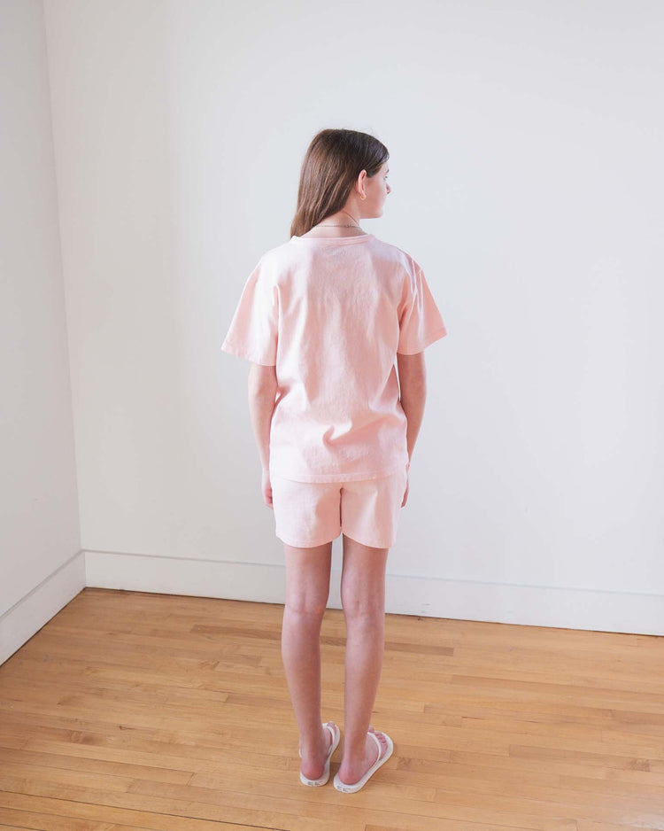 A young person with light brown hair stands on a wooden floor facing away, wearing the Sunchild kazan tee in baby, matching shorts, and slippers in a simple room with white walls.