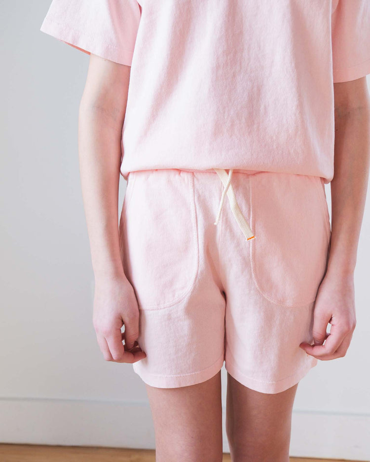 A person models Sunchild merida shorts in baby, a light pink cotton pair with a drawstring waist, and a matching short-sleeve shirt, shown from torso to upper legs against a plain white background.