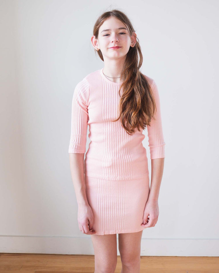 A young girl with long brown hair stands indoors against a white wall, wearing the Sunchild puebla tee in baby. She smiles gently, her arms resting by her sides.