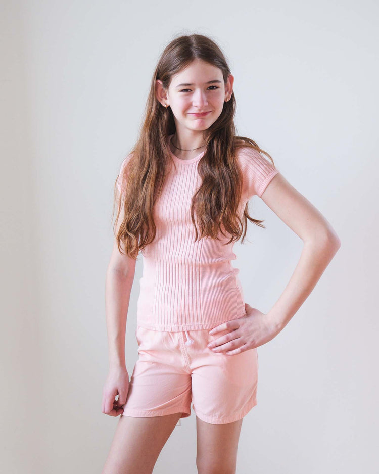A young girl with long brown hair wears Sunchild hunter shorts in baby and a matching light pink top, standing against a white background and smiling with one hand on her hip.