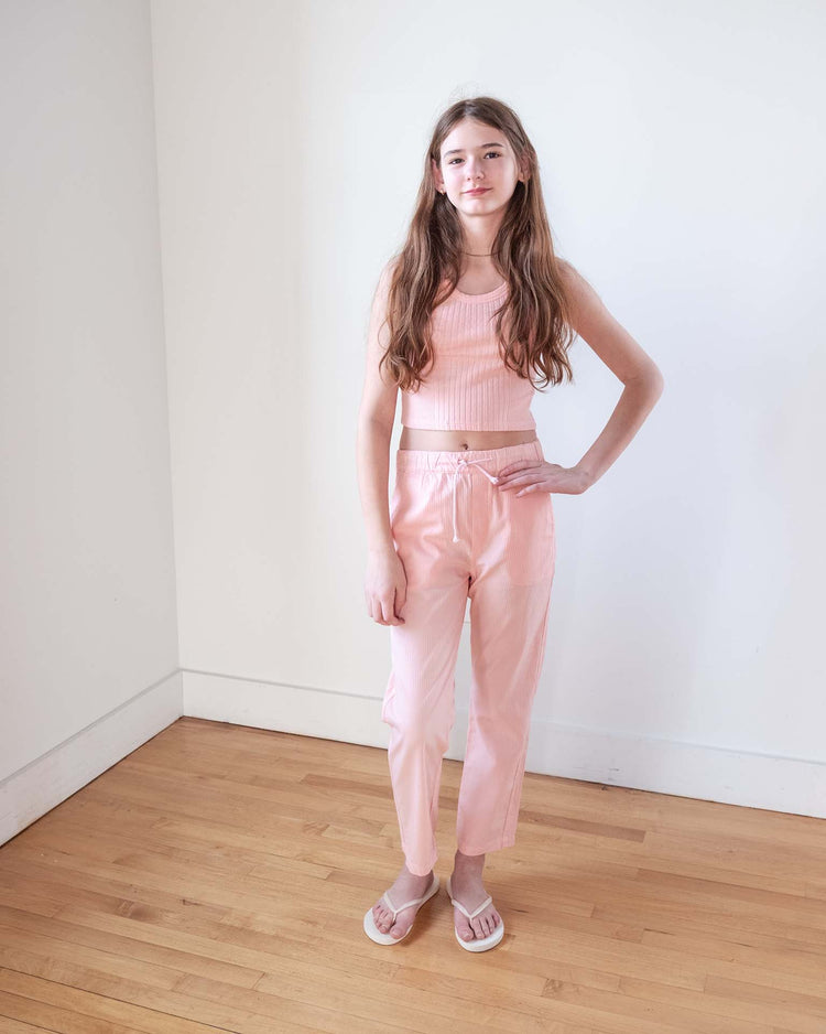 A young person with long brown hair stands in a bright room, wearing the Sunchild cruz tank in baby, matching pink pants, and white slide sandals on a light wood floor.