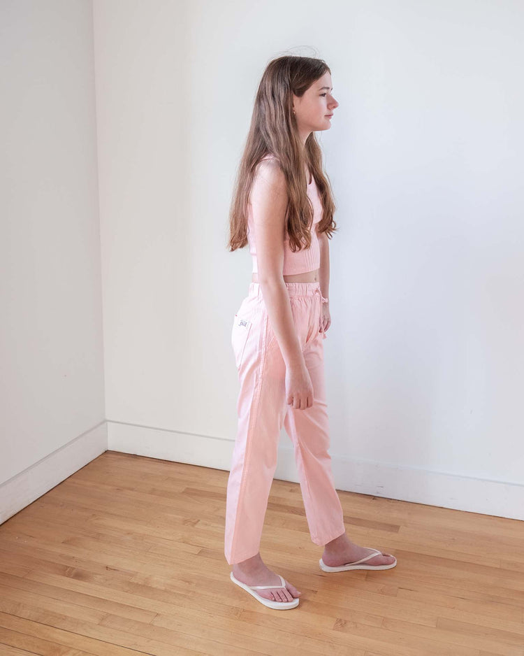 A young woman with long brown hair stands sideways in a room with white walls and a wooden floor, wearing the sunchild cruz tank in baby, pink pants, and white flip-flops.